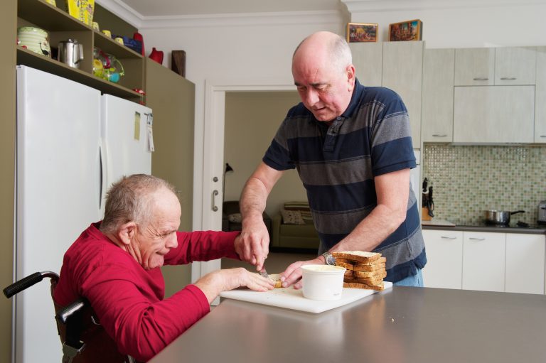 Um homem de meia idade ajuda um homem mais velho com deficiência a passar manteiga no pão