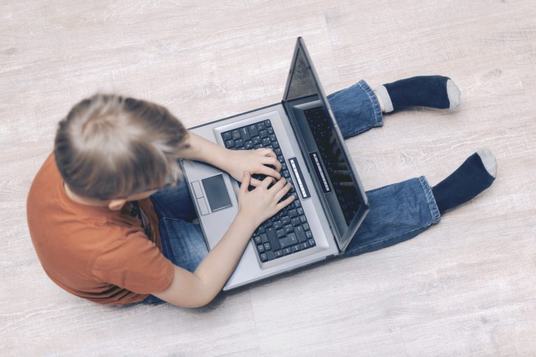 Criança sentada no chão mexendo em um computador que está sobre suas pernas