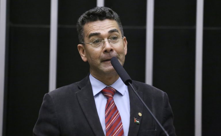 Deputado Padre João fala ao microfone. Ele é claro, tem cabelo escuro, usa óculos, um terno preto, camisa branca e gravata vermelha