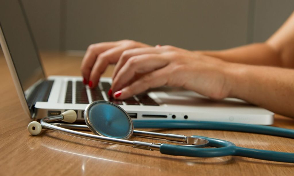 Médica digita no computador, tendo seu estetoscópio ao lado. Foto: National Cancer Institute/Unsplash