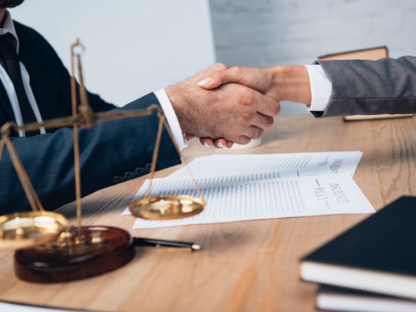 Dois homens apertam as mãos e há uma balança, simbolizando a Justiça, no lado esquerdo