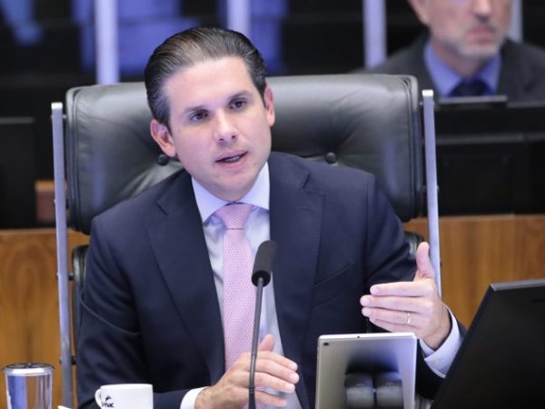Hugo Motta fala ao microfone. Ele é um homem branco, tem o cabelo escuro, veste um terno azul-marinho com camisa branca e gravata vermelha